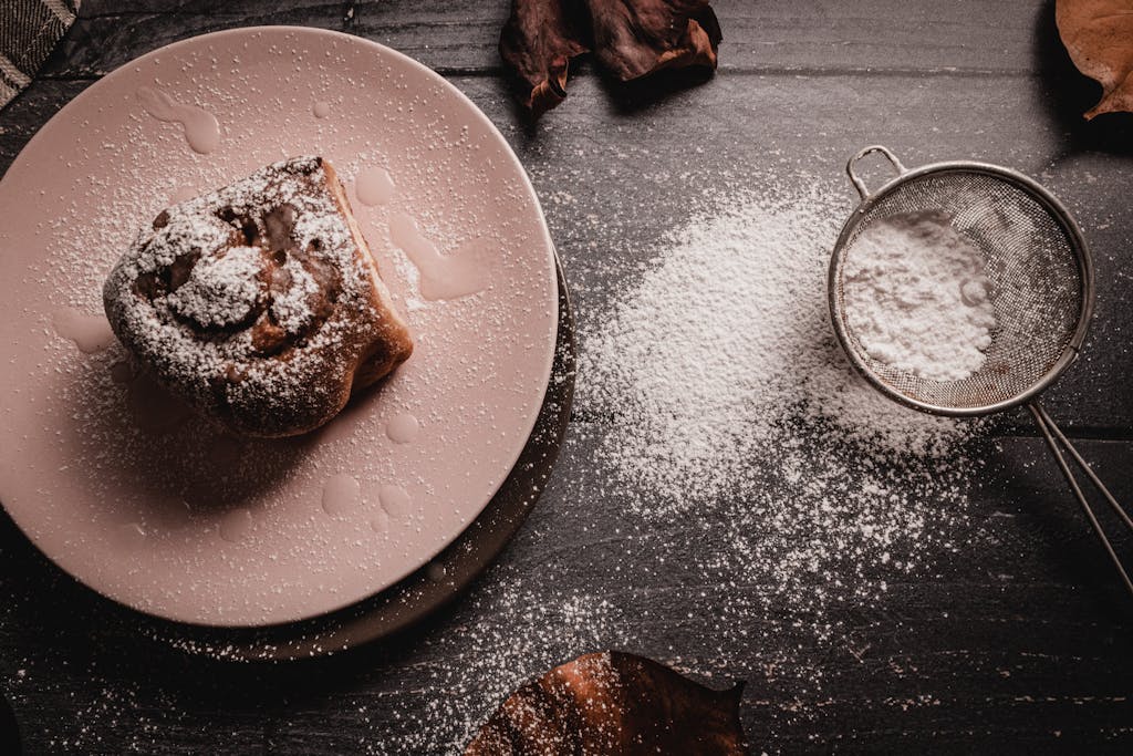 A delicious cinnamon roll dusted with powdered sugar on a pink plate, featuring a sieve on a dark background.