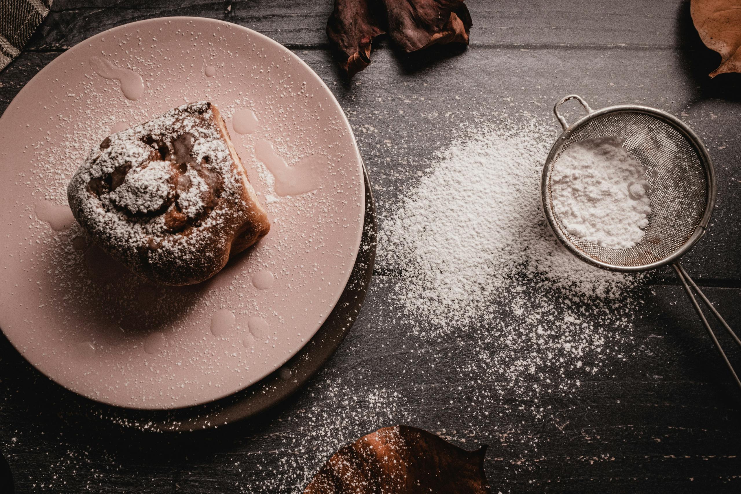 A delicious cinnamon roll dusted with powdered sugar on a pink plate, featuring a sieve on a dark background.