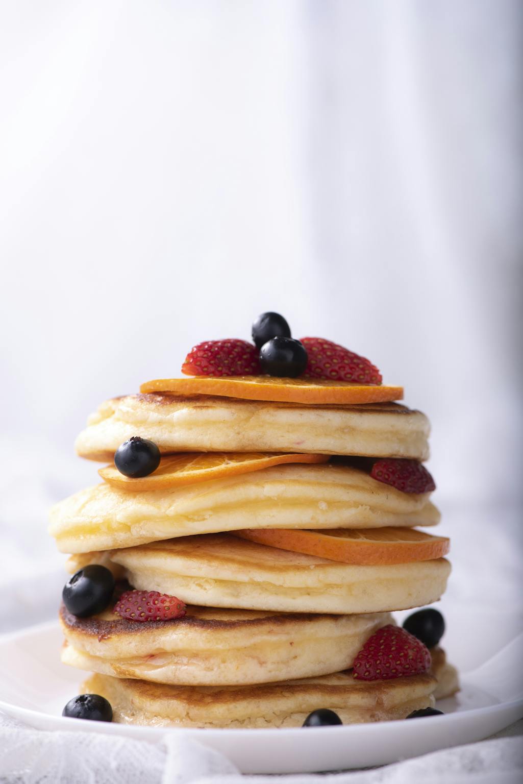 Tall stack of fluffy pancakes topped with blueberries, strawberries, and orange slices.